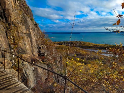 A scenic view of a rocky cliffside overlooking a vibrant blue lake and autumn-colored trees under a cloudy sky.