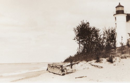 Black and white photo of a lighthouse by the shore, with trees and a sandy beach in the foreground.