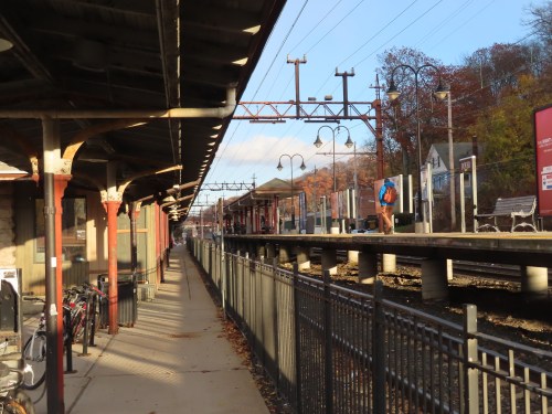 A quiet train station platform with a lone figure, surrounded by autumn foliage and empty bike racks.