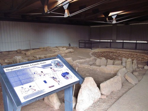 Interior view of a megalithic house site, featuring large stones and an informational sign about its history.