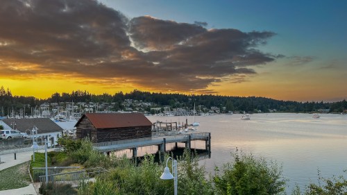Sunset over a marina with boats, a wooden dock, and a rustic building, surrounded by trees and clouds.