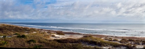 A panoramic view of a serene beach with gentle waves, sandy shore, and a cloudy sky.