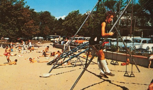 A boy swings on a playground while children play in the sand and cars are parked nearby on a sunny day.