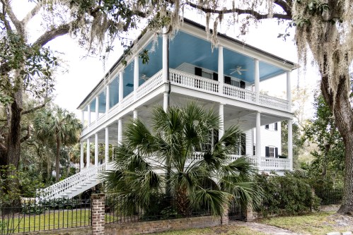 A historic white house with blue accents, featuring multiple porches and surrounded by lush greenery and palm trees.