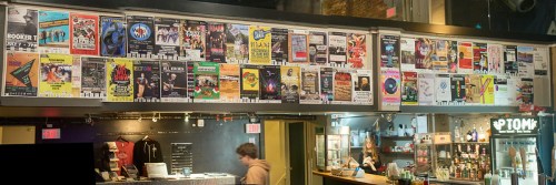 A wall covered in colorful event posters above a bar area, with a person standing in front of the counter.