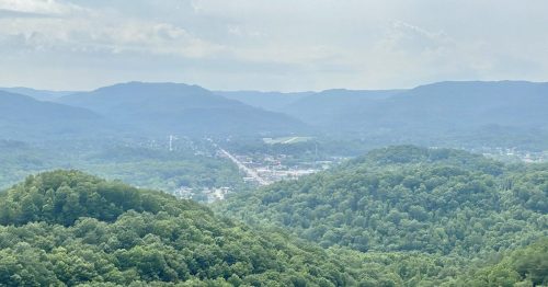 A panoramic view of rolling green hills and a small town nestled in a valley, under a cloudy sky.
