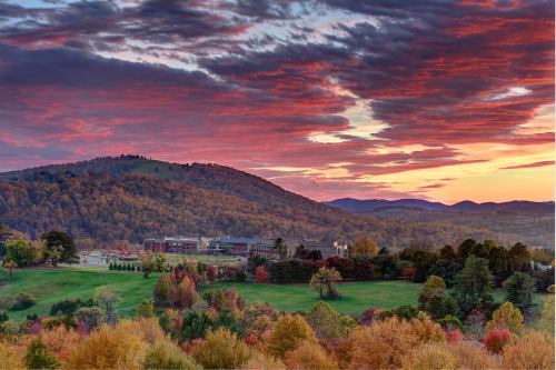 A vibrant sunset over rolling hills, with colorful autumn foliage and a building nestled in the valley below.