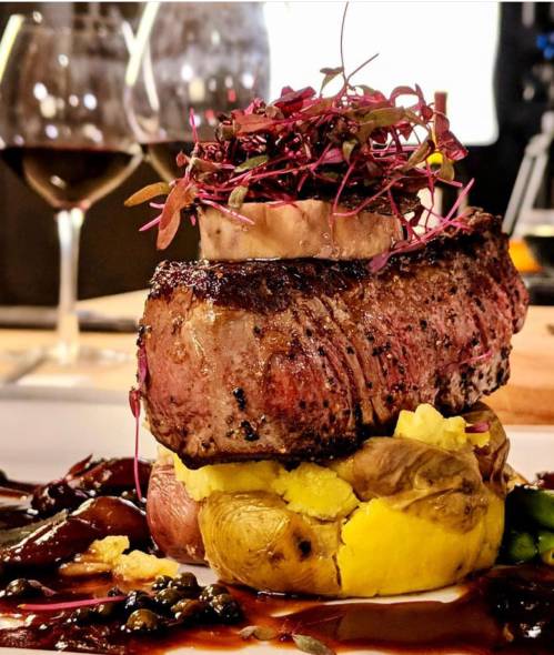 A beautifully plated steak on a bed of potatoes, garnished with microgreens and accompanied by a glass of red wine.