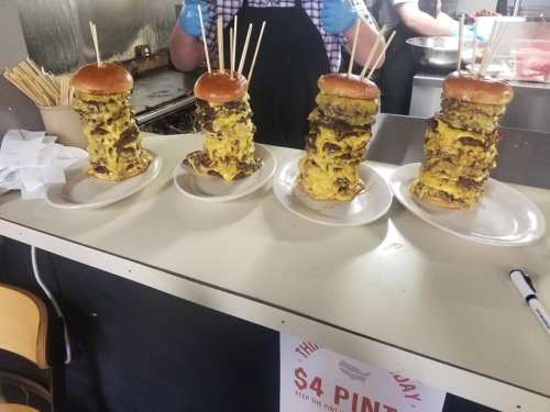 Four towering burgers stacked high with multiple patties and cheese, topped with a bun and skewers, on a counter.