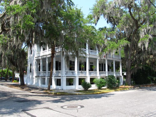 A historic white building with a wraparound porch, surrounded by lush trees and Spanish moss, at a street corner.