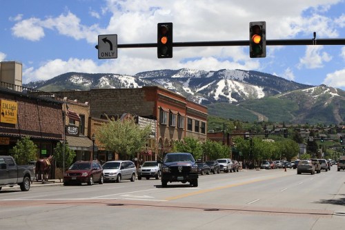A street view of a town with shops, cars, and mountains in the background, featuring snow-capped peaks under a blue sky.