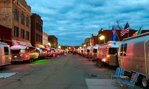 A row of vintage Airstream trailers lined up on a street, illuminated by lights under a cloudy sky.