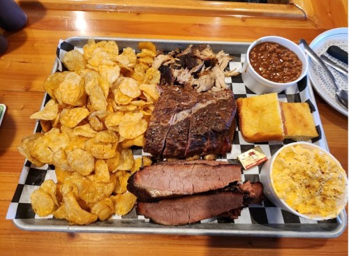 A tray of barbecue featuring brisket, pulled pork, baked beans, cornbread, macaroni and cheese, and potato chips.