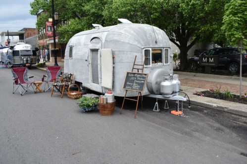A vintage silver trailer with outdoor seating, a sign, and decorative plants, set up on a street for an event.