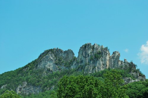 A rugged mountain peak rises against a clear blue sky, surrounded by lush green trees.