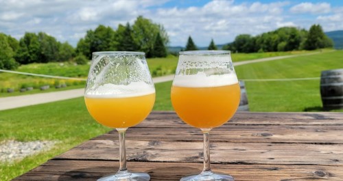 Two glasses of beer sit on a wooden table, with a scenic green landscape and blue sky in the background.