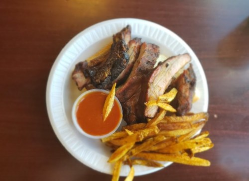 A plate of grilled ribs with a side of fries and a small cup of barbecue sauce.