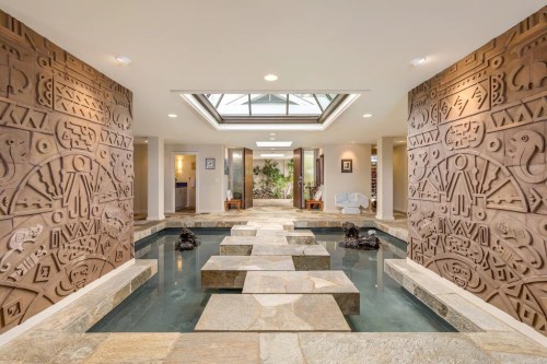 A serene indoor space featuring a stone pathway over water, flanked by intricate textured walls and natural light.