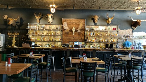 A rustic bar with mounted animal heads, shelves of liquor, and wooden tables in a cozy setting.