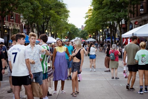 A lively street scene with people socializing and shopping at outdoor stalls under trees.