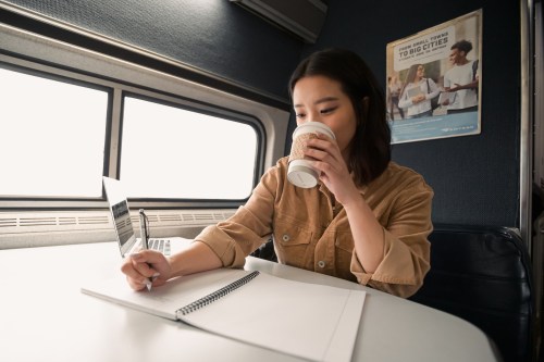 A woman sits at a table, sipping coffee while writing in a notebook, with a laptop open beside her.