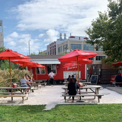 A vibrant outdoor dining area with red umbrellas, picnic tables, and a food truck in a sunny park setting.