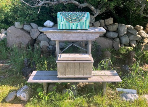 A wooden bench with a decorative box on top, set against a stone wall and surrounded by greenery.