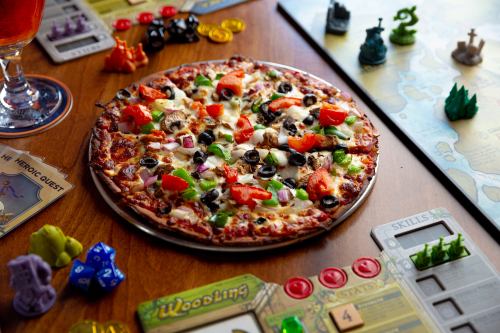A delicious pizza topped with vegetables sits on a table with board game pieces and cards scattered around.