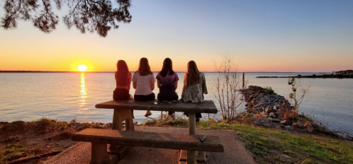 Four people sit on a bench by the water, watching a sunset over the horizon.
