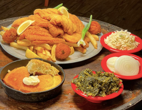 A platter of fried fish with fries, lemon, and hushpuppies, alongside sides of cornbread, collard greens, and coleslaw.