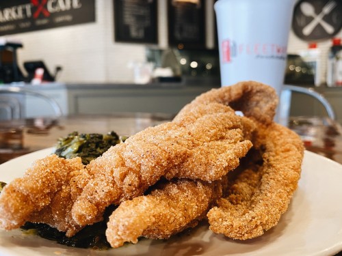 A plate of golden-brown fried catfish fillets with a side of greens and a drink in the background.