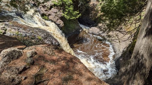 A scenic view of a waterfall cascading into a rocky pool, surrounded by trees and pine needles on the ground.