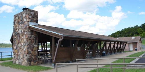 A wooden pavilion with a stone chimney by a lake, surrounded by green grass and trees under a blue sky.