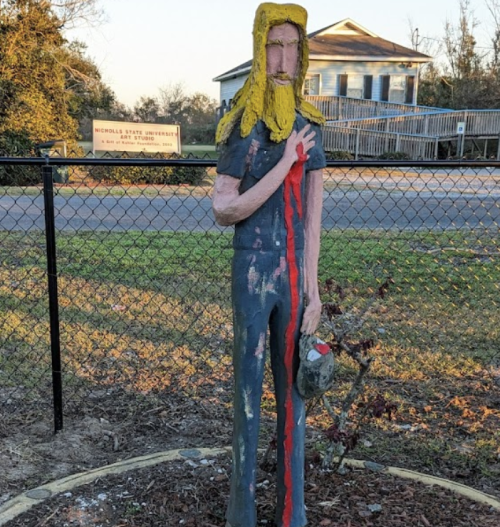 A painted wooden statue of a person with long hair, holding a helmet and standing in a garden near a fence.