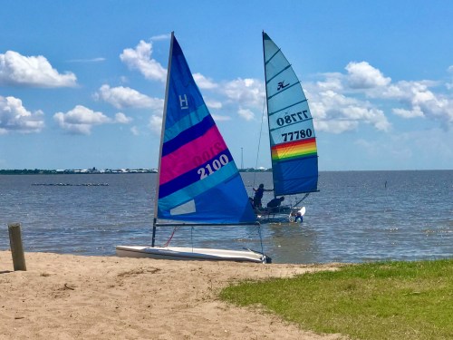 Two sailboats with colorful sails on a sandy beach by a calm lake under a blue sky with fluffy clouds.
