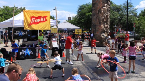 Children playing with hula hoops at a lively outdoor event, with tents and a sign for French's in the background.