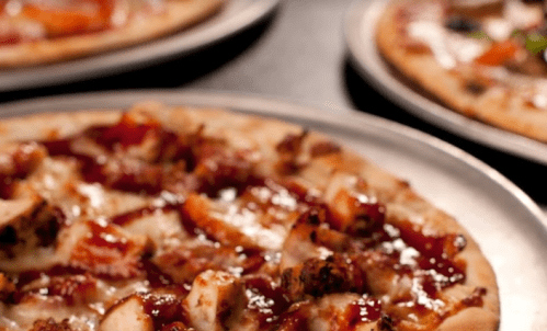 A close-up of a pizza topped with chicken and barbecue sauce, with other pizzas blurred in the background.