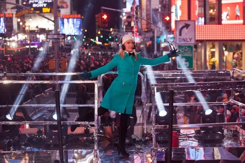 A woman in a teal coat and white hat stands on a stage in Times Square, surrounded by bright lights and a large crowd.