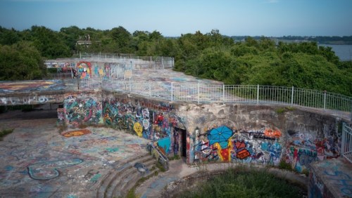 Abandoned structure covered in colorful graffiti, surrounded by lush greenery and overlooking a body of water.