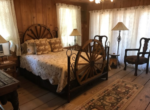 Cozy rustic bedroom with a wooden bed, decorative pillows, two lamps, and large windows with sheer curtains.