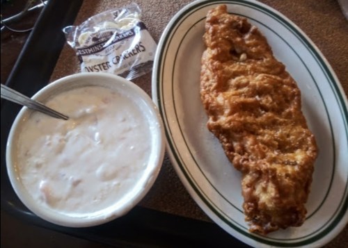 A plate with a large fried fish fillet, a bowl of clam chowder, and a packet of oyster crackers on the side.