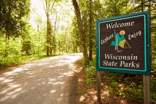 A sign welcoming visitors to Wisconsin State Parks, surrounded by lush green trees along a gravel road.