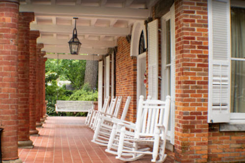 A porch with white rocking chairs and brick columns, surrounded by greenery and natural light.
