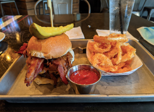 A bacon sandwich with a pickle on top, served with crispy onion rings and a side of ketchup on a metal tray.