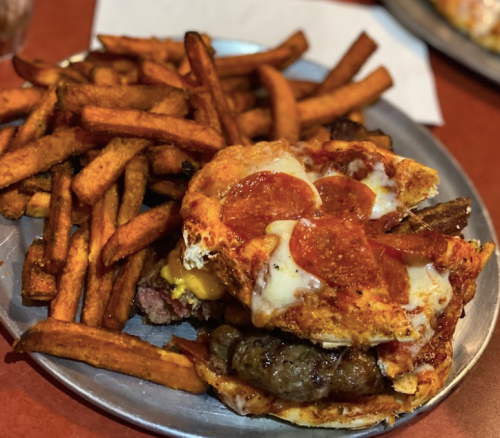 A plate with a stacked burger topped with pepperoni and cheese, served with a side of crispy sweet potato fries.