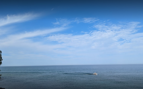 A calm sea under a blue sky with wispy clouds, featuring a small boat leaving a wake in the water.