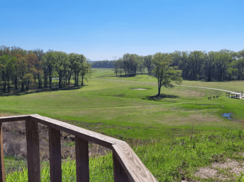 A scenic view of a green landscape with trees, a clear blue sky, and a wooden railing in the foreground.