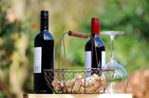 A wire basket filled with wine corks, with two bottles of red wine and a wine glass in the background.