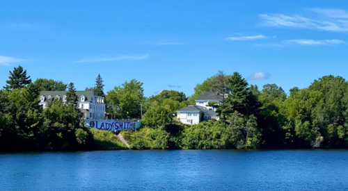 A serene lake view featuring two buildings surrounded by lush greenery under a clear blue sky.