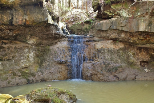 A small waterfall cascades over rocky cliffs into a calm, greenish pool surrounded by trees.